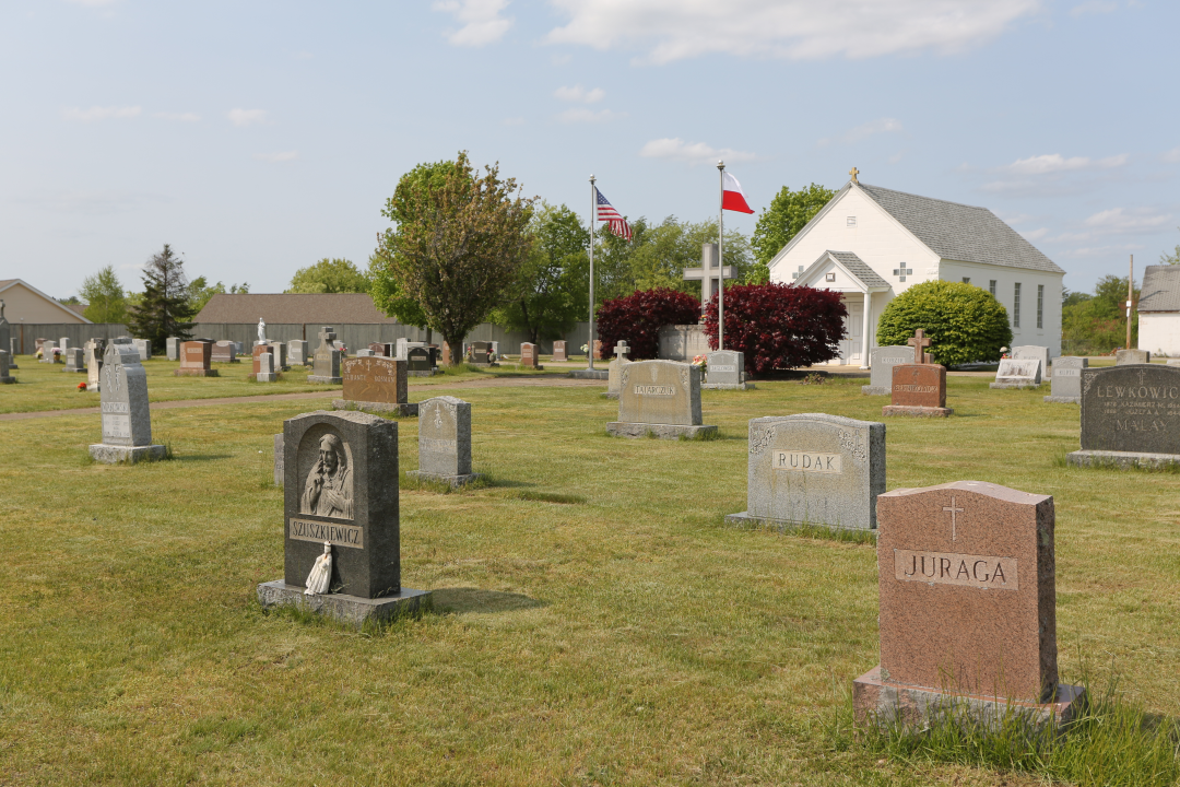 Widok cmentarza z nagrobkami stojącymi na zielonej trawie w tle kościół i flagi