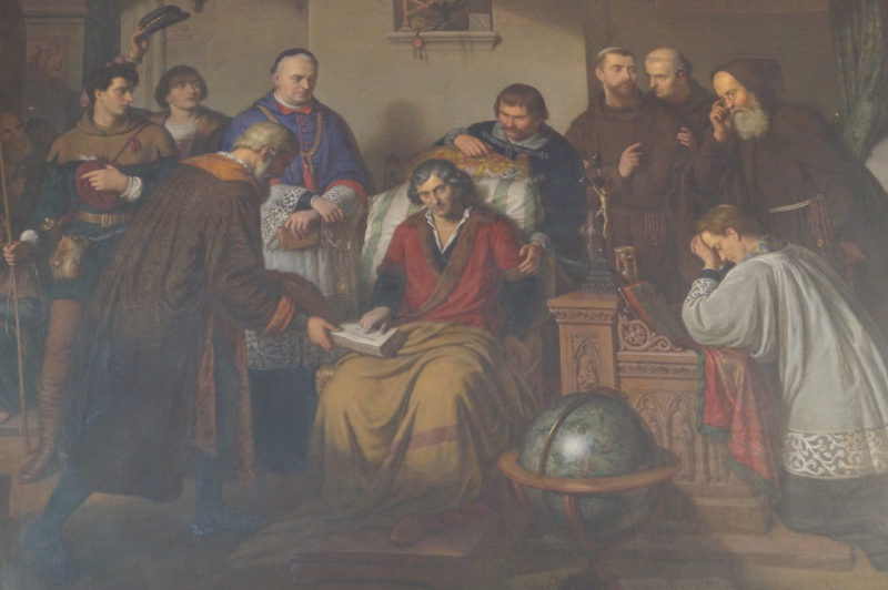 Obraz przedstawiający siedzącego z kocem na kolanach Mikołaja Kopernika obok dwóch księży